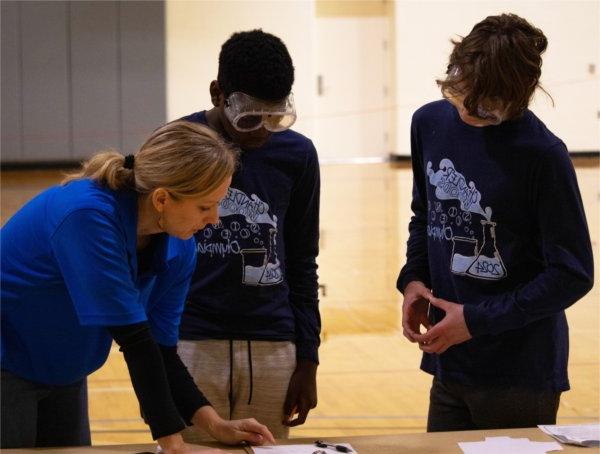 两个学生戴着护目镜看着桌子，右边是一个穿着蓝色衬衫的教练