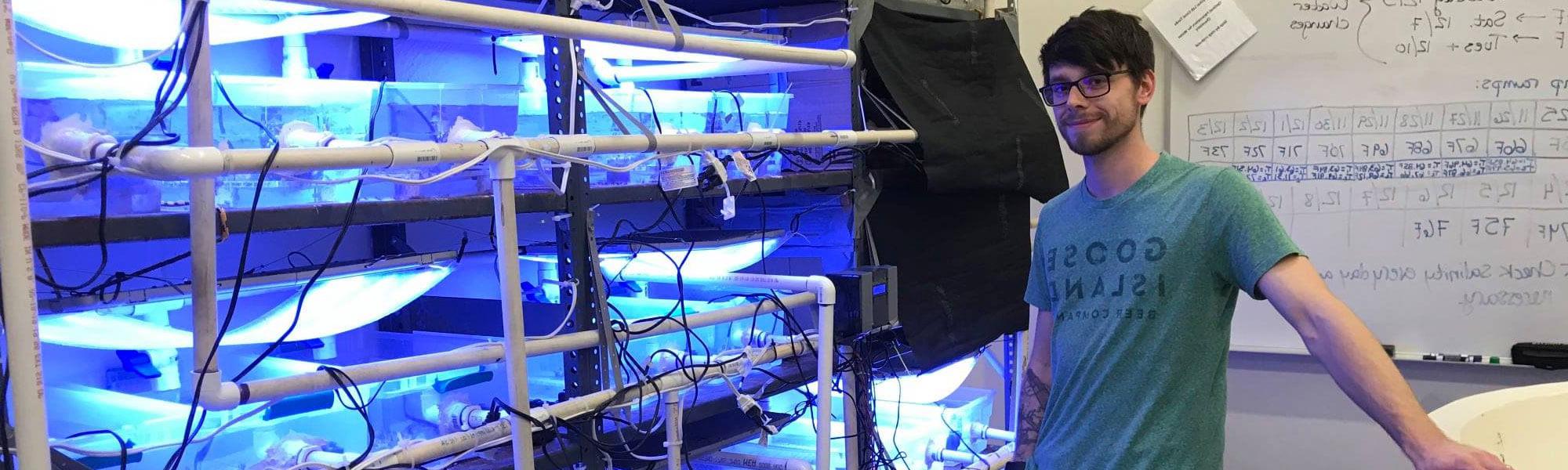 MS生物学研究生Tyler Harman在马斯基根的Annis水资源研究所进行冷水珊瑚研究，并在定制的循环水族馆系统前摆姿势.